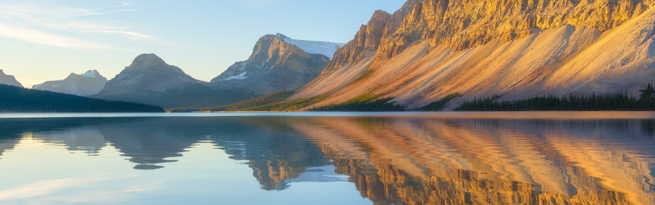 A mountain landscape during sunrise. Mountains stand against a clear sky, reflected in the still surface of a lake.