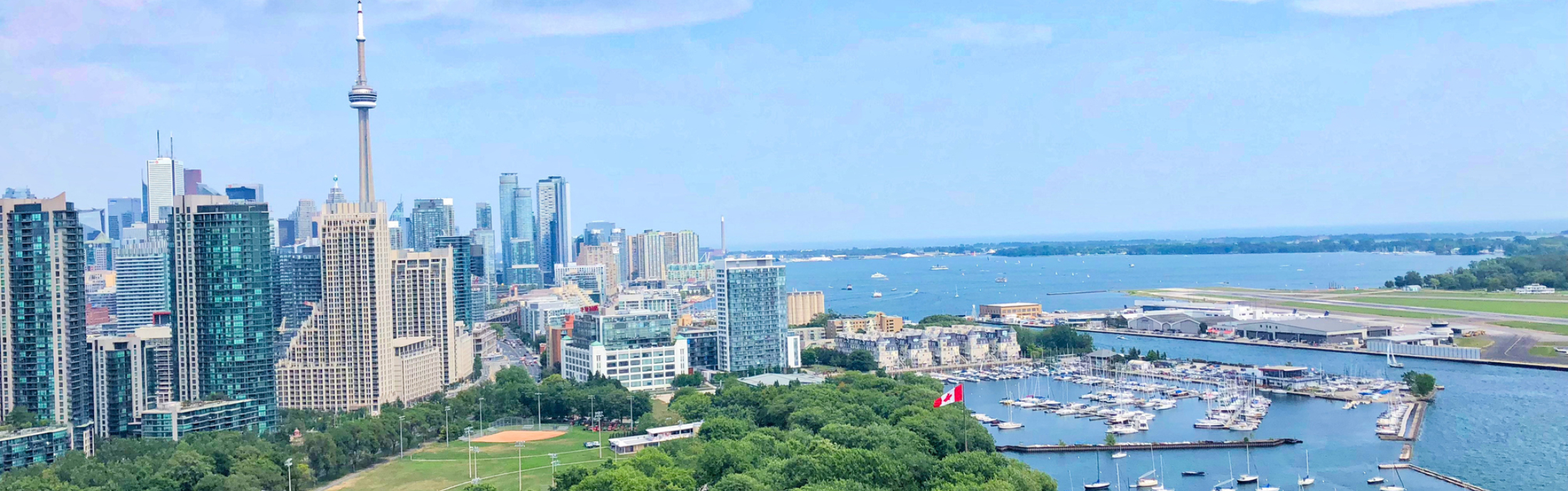 Skyline of Toronto with the CN Tower and skyscrapers. In the foreground, a green park and marina with sailboats.