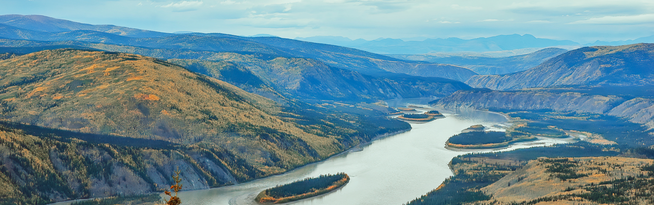 Aerial view of a winding river surrounded by forested mountains and a cloudy sky above.