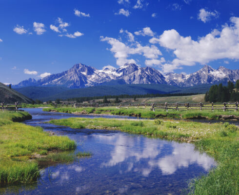 A serene landscape with a clear stream, green grass, and a wooden fence. Snow-capped mountains are in the distance.