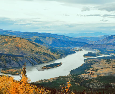 Aerial view of a winding river surrounded by forested mountains and a cloudy sky above.