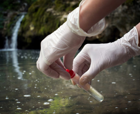 Two gloved hands holding a test tube above a body of water. 
