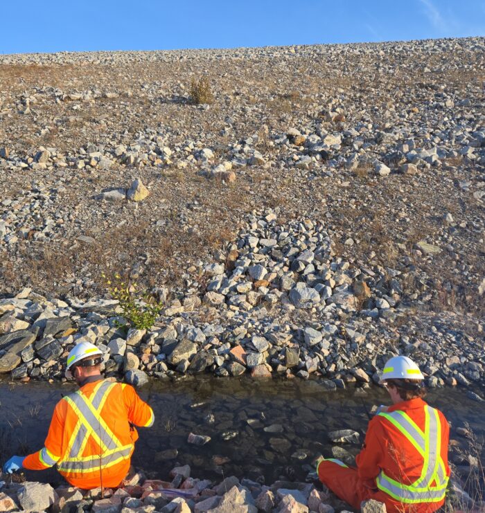 Duncan McDonald, Environmental Scientist, and Mike Bower, Environmental Scientist, wearing Hi-Vis jackets, collecting a water sample from a stream.
