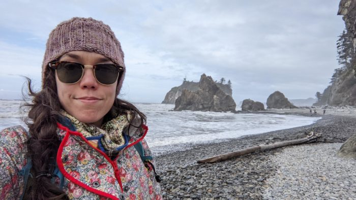 Mackenzie Bromstad standing on a pebble beach with the ocean and various ocean rocks stretching behind her.