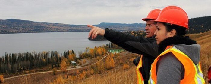 Two Ecometrix team members in orange safety vests and hard hats stand on a hillside with a lake in the background. One points off camera.